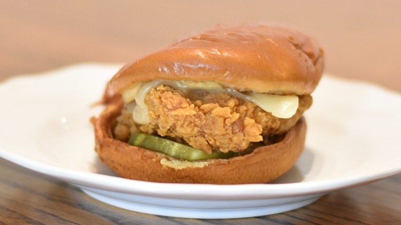 A Popeyes chicken sandwich with golden barbecue sauce, cheese, and pickle, on a white plate