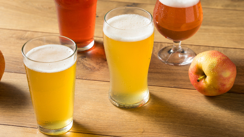 Various pints of hard apple cider sit on a wooden table with an apple