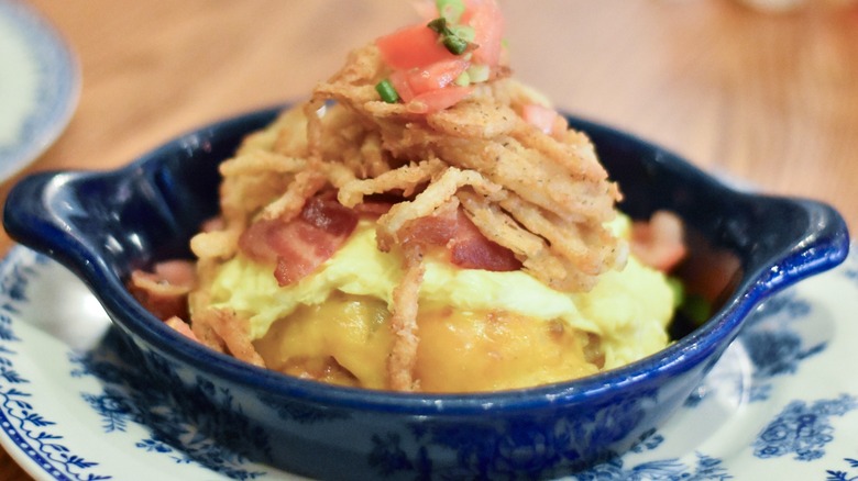 A dark blue dish of hashbrown casserole with eggs, bacon, fried onions, and diced tomatoes