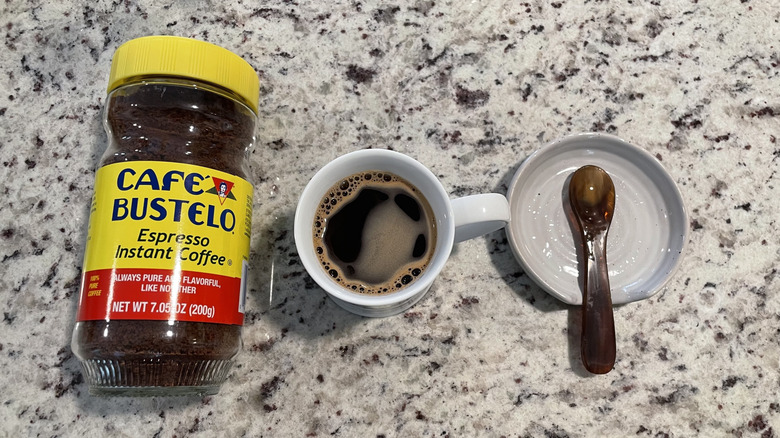 Cafe Bustelo coffee packaging is next to a cup of coffee and a spoon on a saucer.