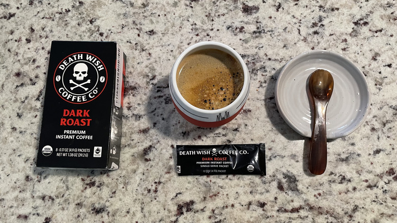 Death Wish coffee packaging is next to a cup of coffee and a spoon on a saucer.