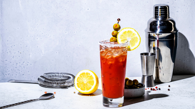 A Bloody Mary in a tall glass with olives in front of a cocktail shaker and strainer