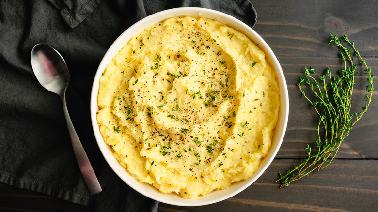 A bowl of creamy garlic mashed potatoes, topped with chopped thyme and ground pepper over a black tea towel on a brown, wooden table, a spoon on one side, a sprig of thyme on the other
