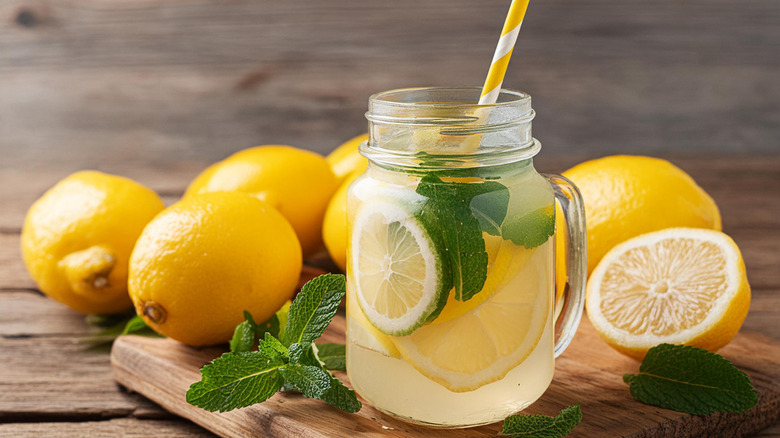 A mason jar glass of lemonade on a wooden board surrounded by mint and lemons.