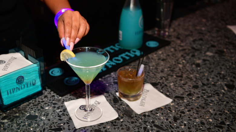 A bartender places a glowstick inside a blue cocktail in a martini glass with a Hpnotiq bottle in the background