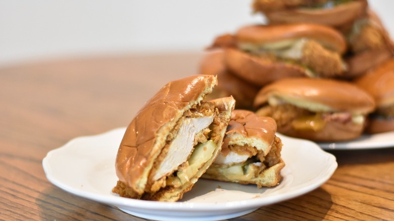 A Popeyes chicken sandwich with pickles cut in half on a white plate with other sandwiches stacked in background