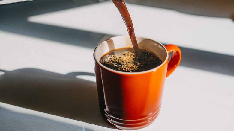 A metal carafe pour black coffee into an orange coffee mug