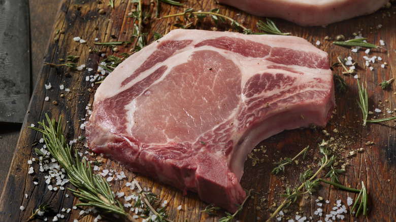 Two raw pork chops on a wooden board surrounded by salt and fresh herbs