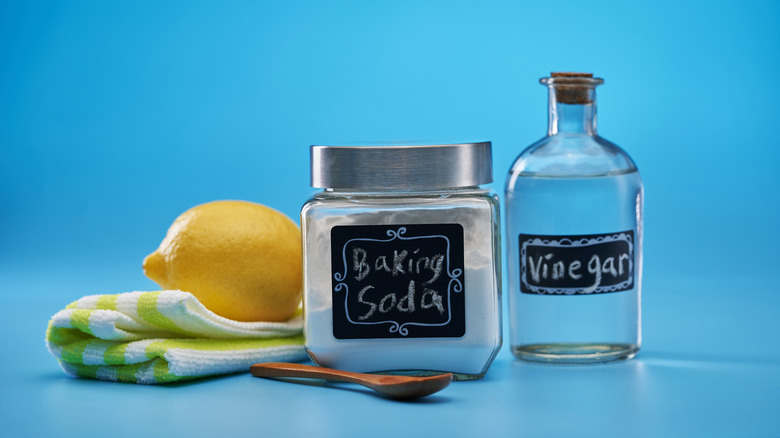 Bottles of baking soda and vinegar with handmade chalk labels sit next to a lemon on a yellow and white striped dish towel with a wooden spoon in front on a blue background.