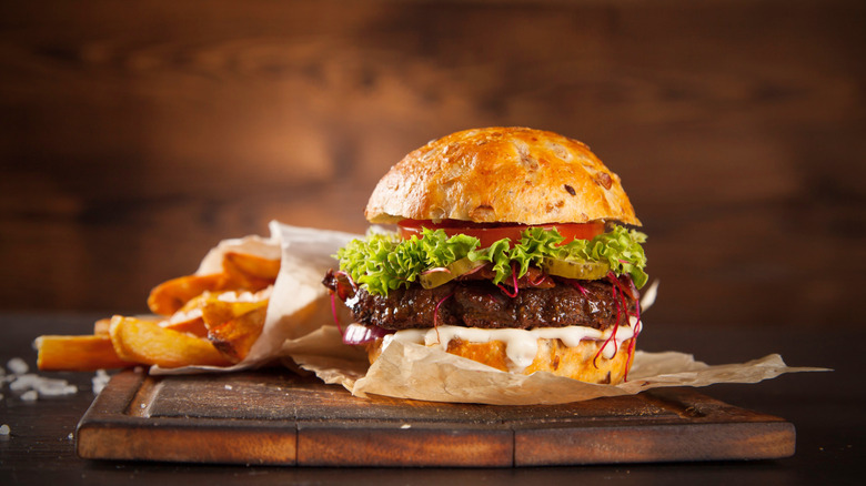A burger with various condiments sitting on a cutting board next to french fries.