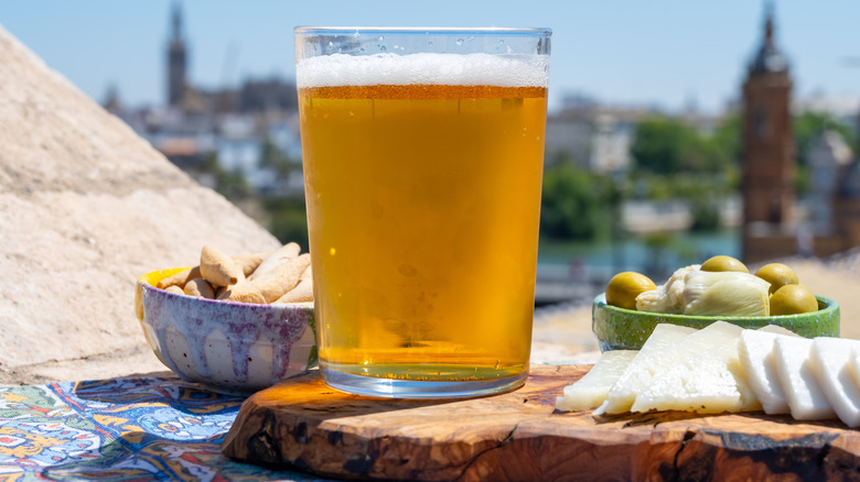 Glass of beer on wood board with cheese surrounded by bowls of appetizers and Seville, Spain, in background