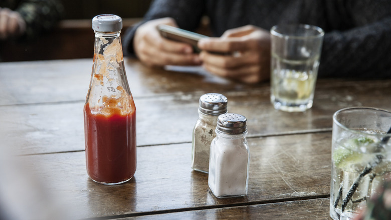 A glass ketchup bottle on a table with salt and pepper.