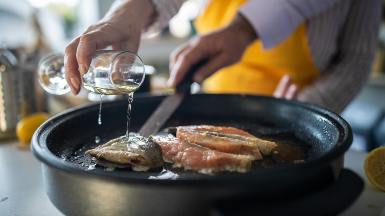 A person's hand pours wine into a pan of fish on the stove.