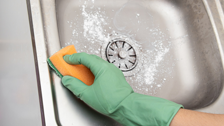 Hand with green glove scrubbing kitchen sink with sponge