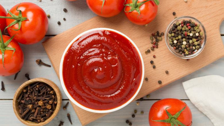 small white bowl of ketchup on cutting board beside fresh tomatoes and peppercorns