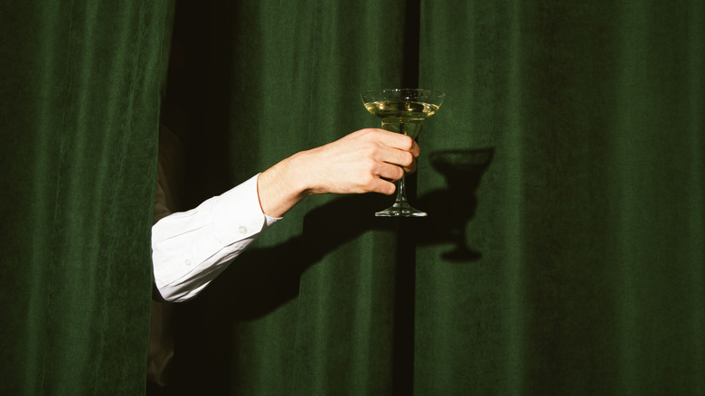 arm holding a martini cocktail from behind a green curtain