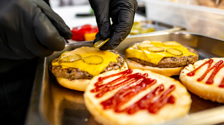 A pair of black gloved hands placing pickle slices on a cheeseburger.