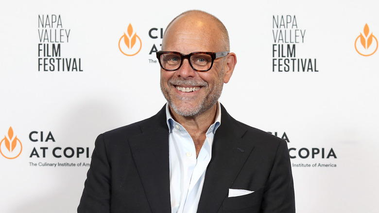 Alton Brown smiles in a black suit and glasses at a red carpet event