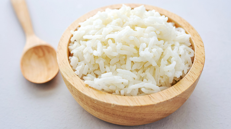 A wooden bowl of white rice on a grey countertop with a wooden spoon next to it.
