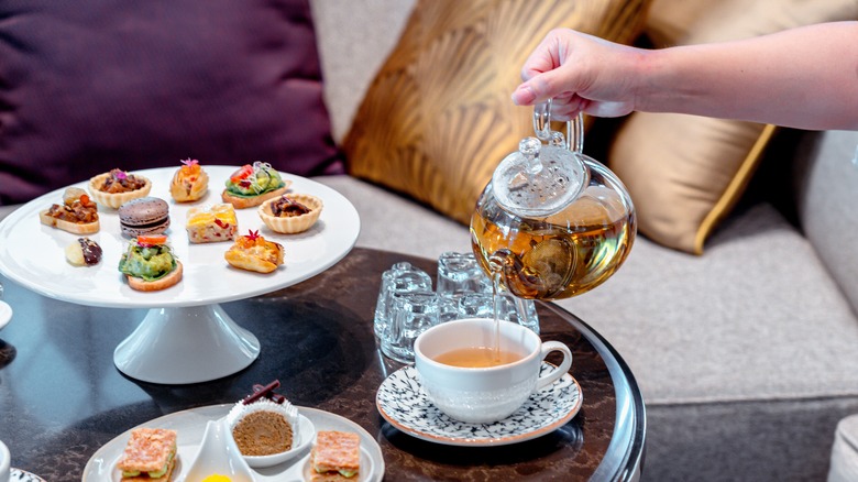 English afternoon tea set including hot tea, pastry, scones, sandwiches, and mini pies on marble top table.
