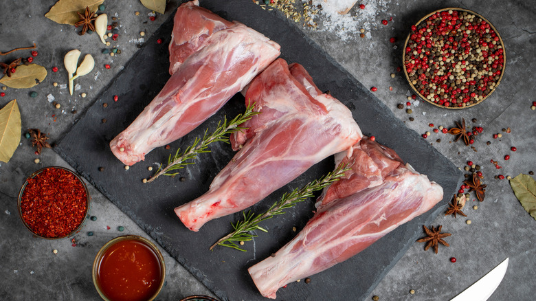 Three raw lamb shanks on a stone slate with rosemary, surrounded by bowls of dry spices