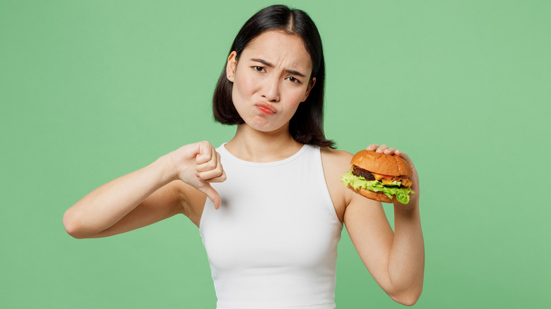 Woman holding fast food burger and giving thumbs down gesture