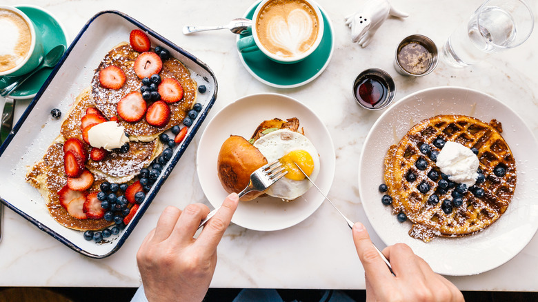 Person cutting into a fried egg burger surrounded by a breakfast spread of pancakes, waffles, and coffee