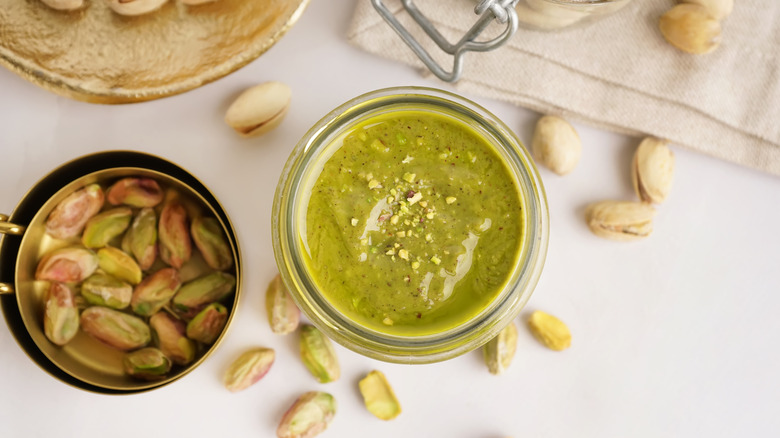 A jar of pistachio butter with shelled pistachios around it on a white counter
