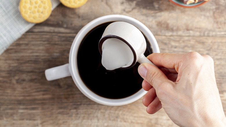 A hand pouring creamer into a cup of coffee over a rustic wooden countertop, a couple of cookies on a tea towel above it.