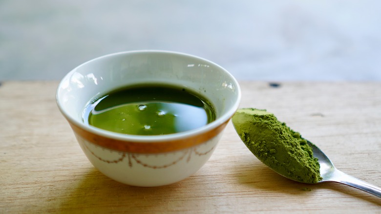 a spoon of ground matcha powder pictured alongside a bowl of brewed matcha