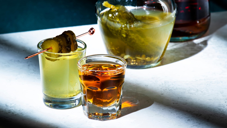 A pickleback shot setup, with a shot glass of whiskey, another shot glass of pickle juice, garnished with pickle slices, and a jar or pickles and bottle of whiskey in the background.