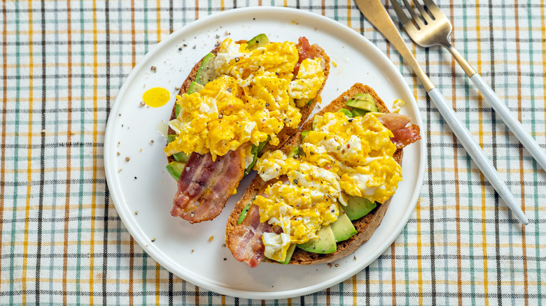avocado toast topped with bacon and scrambled eggs on a white plate