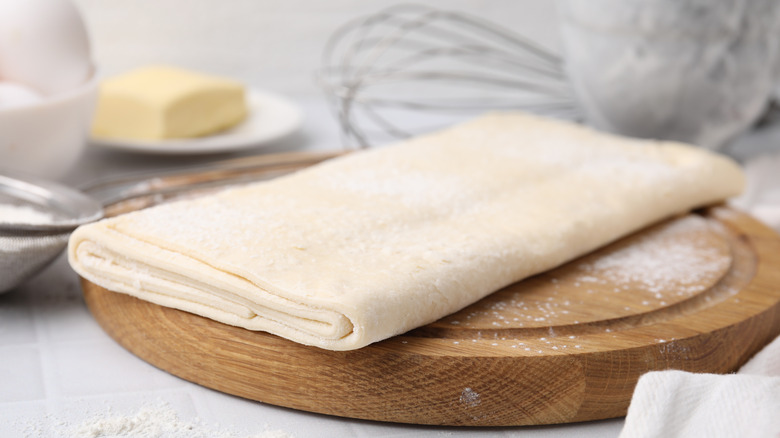 Uncooked puff pastry is placed on a wooden cutting board; baking tools are in the background.