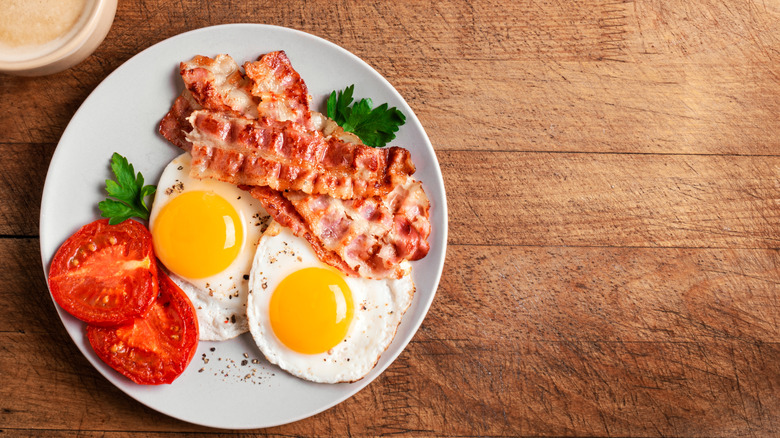 A breakfast plate of eggs, bacon, and tomato slices on a wood table