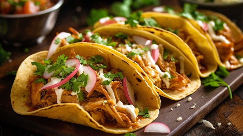 Four pork tacos in a row on a wooden serving tray, topped with pickled onions, cilantro, and crema.
