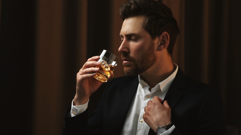 a well-dressed gentleman having a sip of whiskey on the rocks in front of a brown curtain