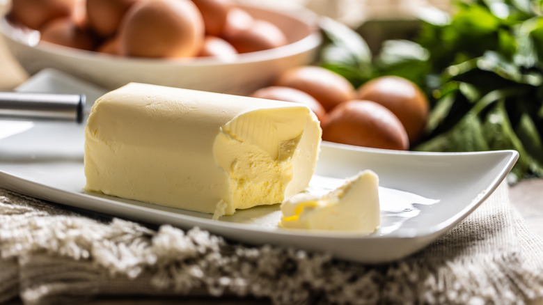 A portion of butter has been sliced into on a plate with brown eggs out of focus in the background.
