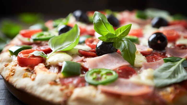 A thin crust pizza with tomatoes, ham, olives, and fresh basil