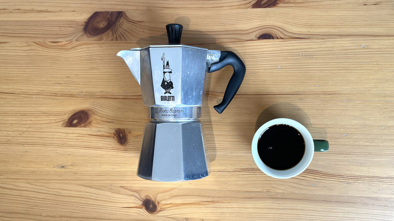 A moka pot lies flat next to a cup of coffee shown from above