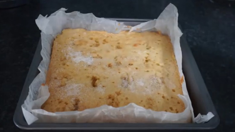 A lemon cake partially covered with sugar, nestled in parchment paper in a metal baking pan