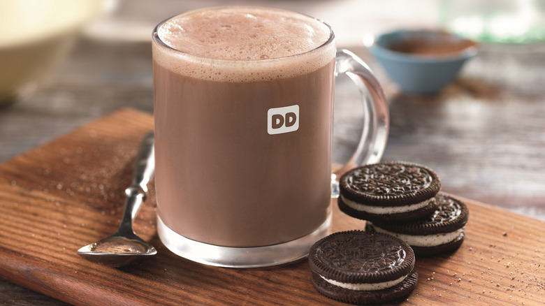 A clear glass mug of hot chocolate on a cutting board with a spoon and three Oreo cookies
