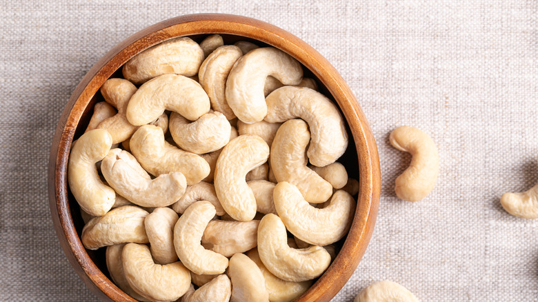 A brown bowl of cashews on a woven tablecloth with cashews around it