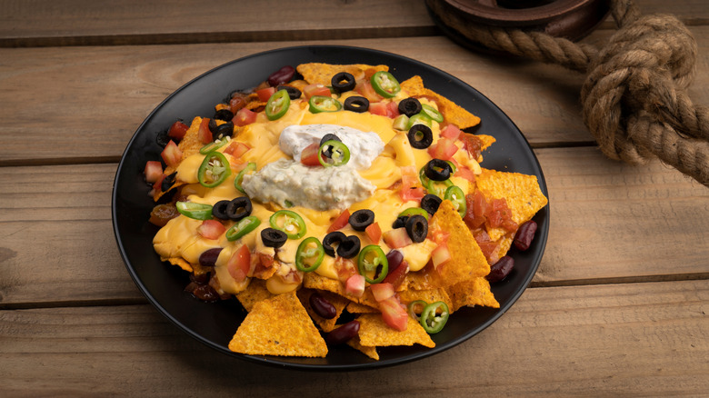 A plate of nachos covered in cheese sauce, tomatoes, olives, beans, jalapenos, and sour cream