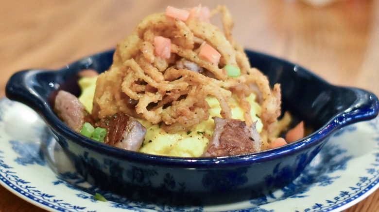 A dark blue dish of hashbrown casserole with steak, eggs, and fried onions