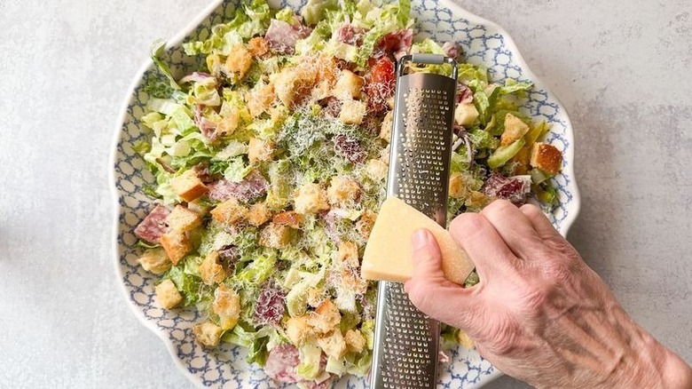 Grating parmesan onto Italian deli grinder salad in large serving bowl