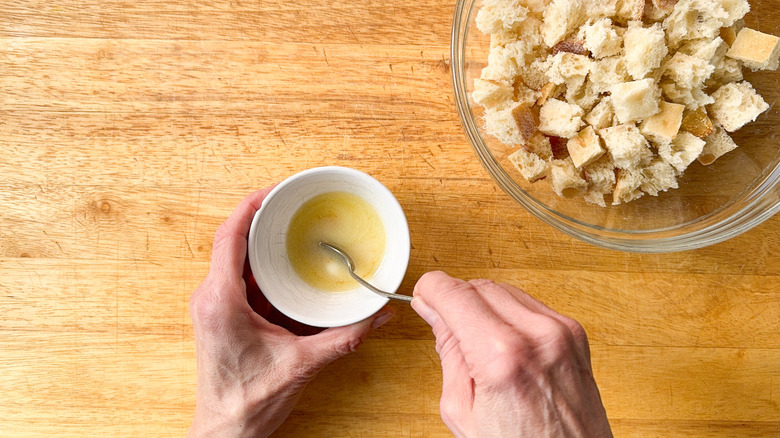 Stirring melted butter and garlic powder in small bowl next to glass bowl of bread cubes