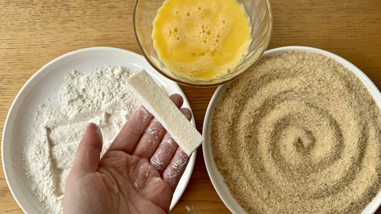 hand holding a cheese stick above three white bowls filled with breadcrumbs, flour, and eggs