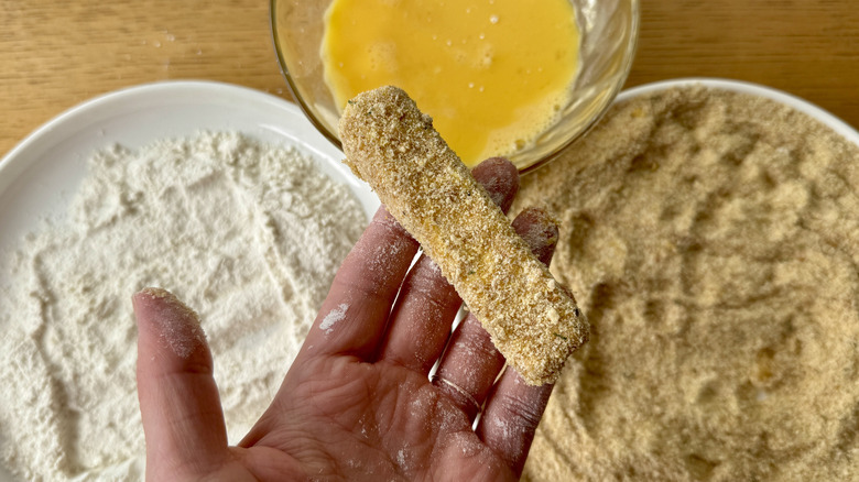 hand holding a breaded cheese stick above three white bowls filled with breadcrumbs, flour, and eggs