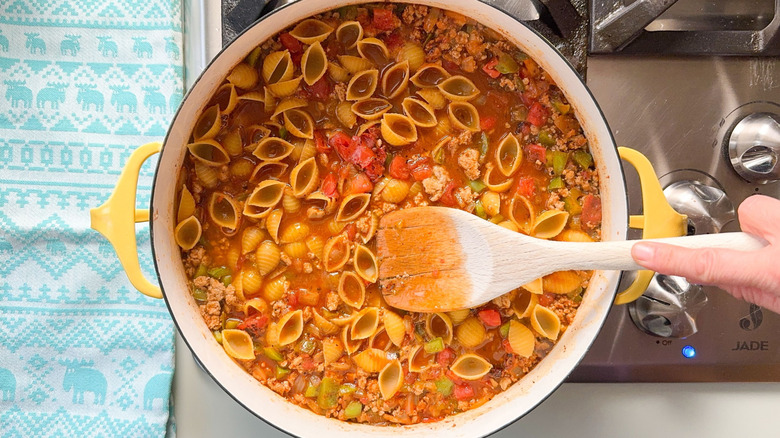 Stirring shell pasta into ground turkey, tomatoes, and broth in pot with wooden spoon
