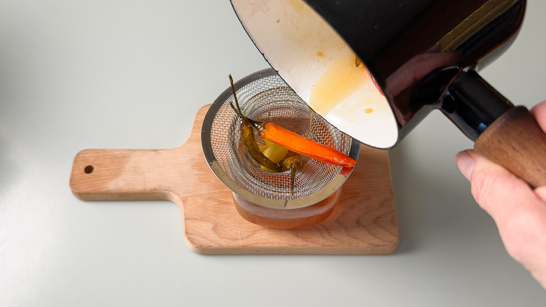 Pouring hot honey through strainer with hot chiles over jar on cutting board